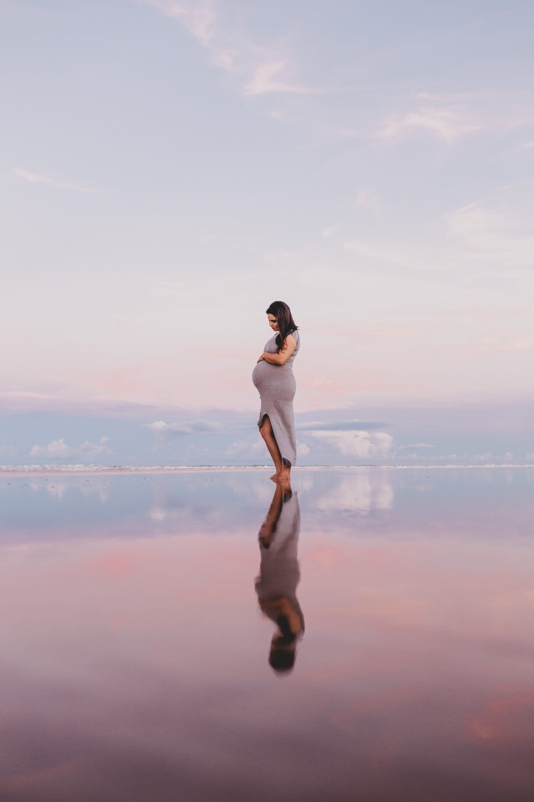 A pregnant woman is standing by a lake at sunset.