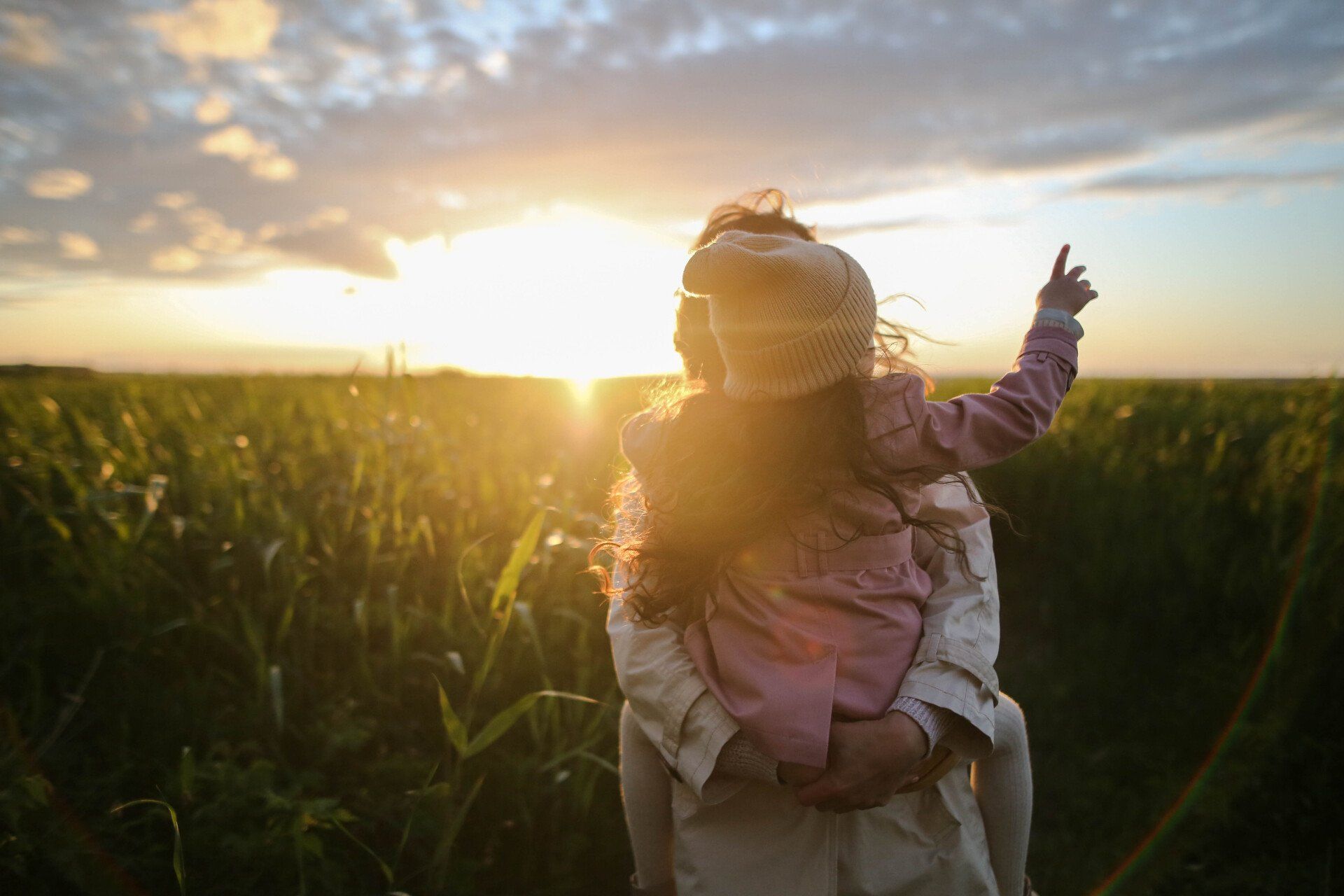 A mom holding her toddler standing in a cornfield at sunset.